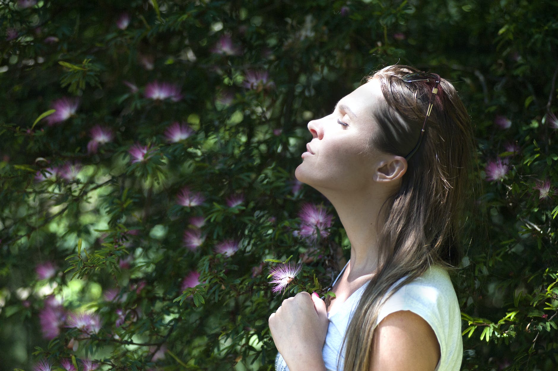 woman closing her eyes against sun light standing near purple petaled flower plant