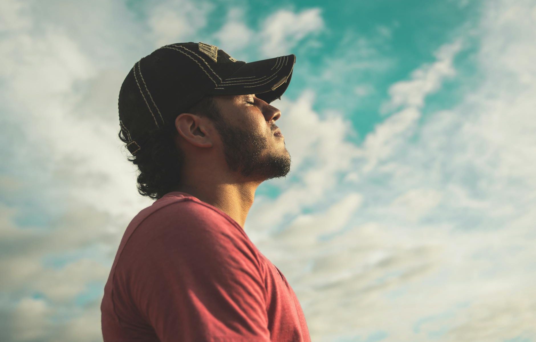 man wearing black cap with eyes closed under cloudy sky